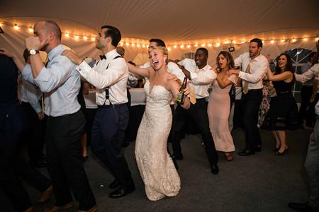 A bride and wedding guests form a lively conga line and dance together under string lights at a reception, energized by vibrant wedding music performed by a talented Boston band.