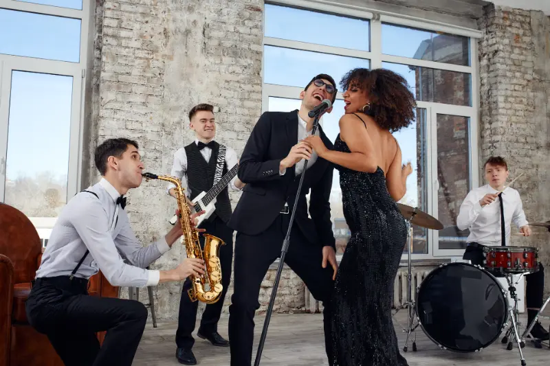 A Boston Band performs indoors; a man plays saxophone, another plays electric guitar, a third plays drums, while a man and woman sing into a microphone together—perfect for unforgettable wedding music.