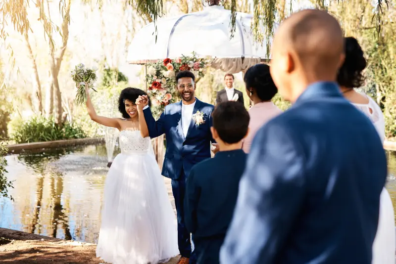 A bride and groom stand together smiling and waving at guests during an outdoor wedding ceremony near a pond, while Boston Band plays enchanting wedding music in the background.